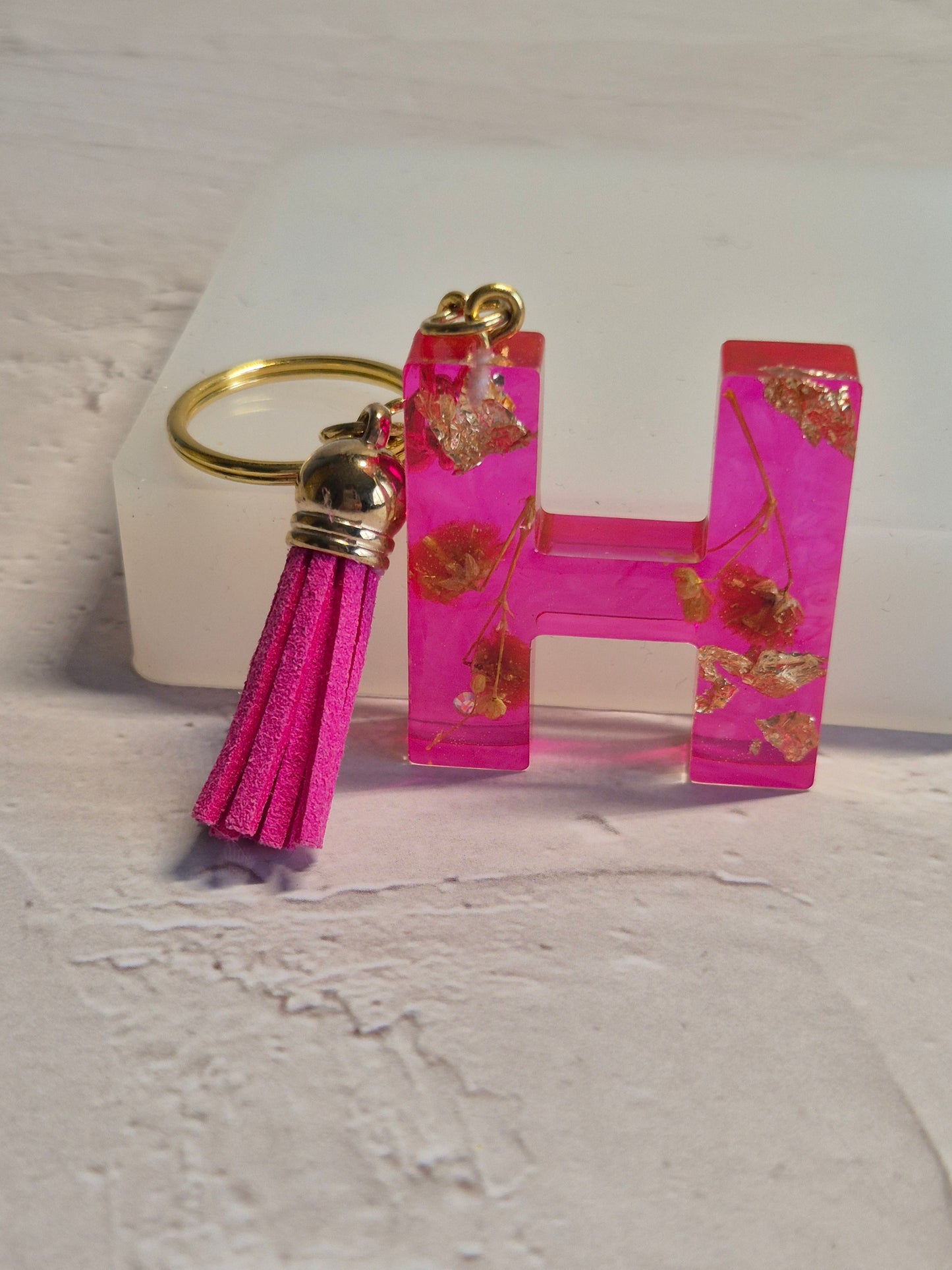 Llavero letra H de resina epoxi fucsia transparente con flores secas, pan de oro y borla fucsia