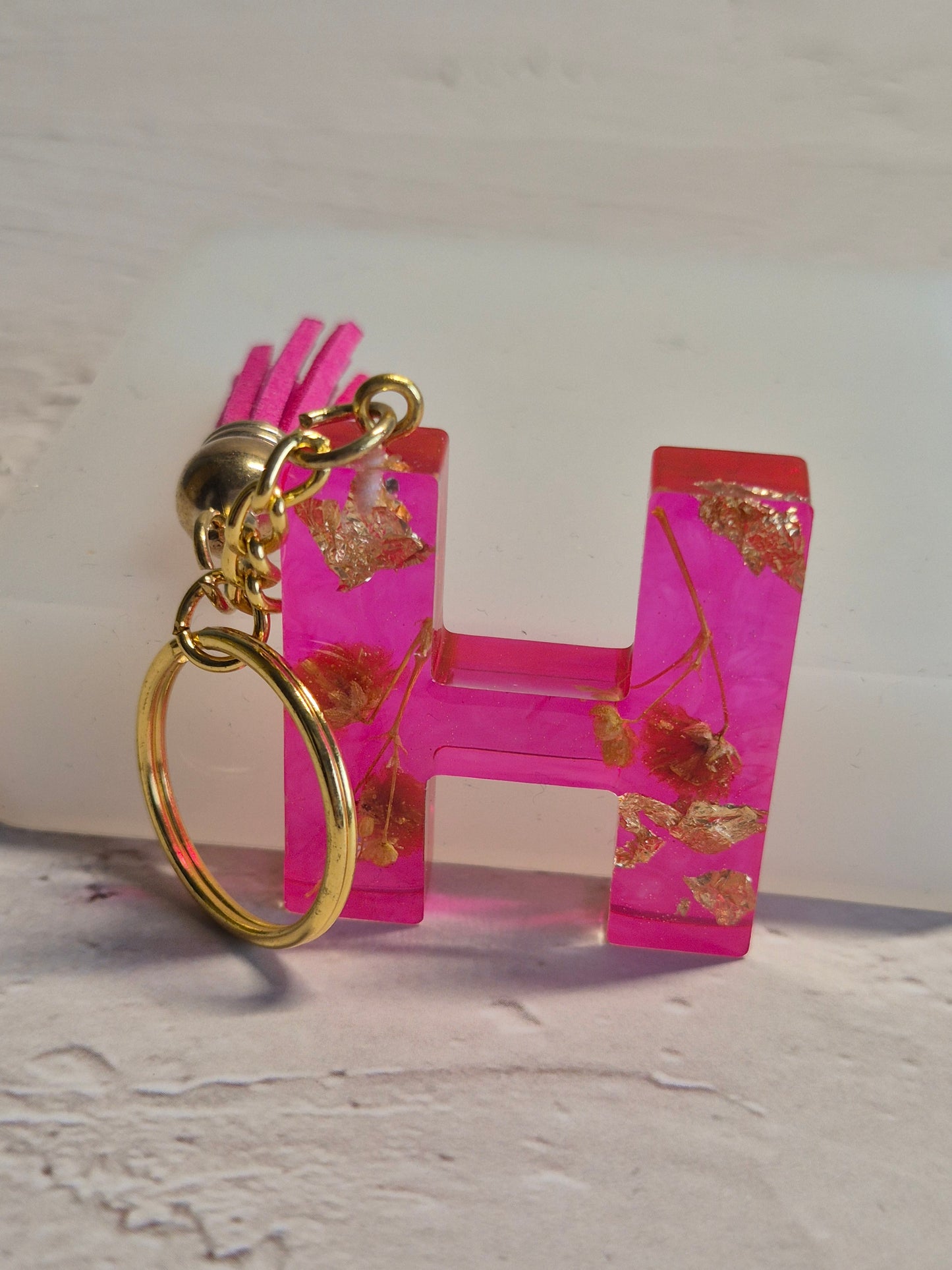 Llavero letra H de resina epoxi fucsia transparente con flores secas, pan de oro y borla fucsia