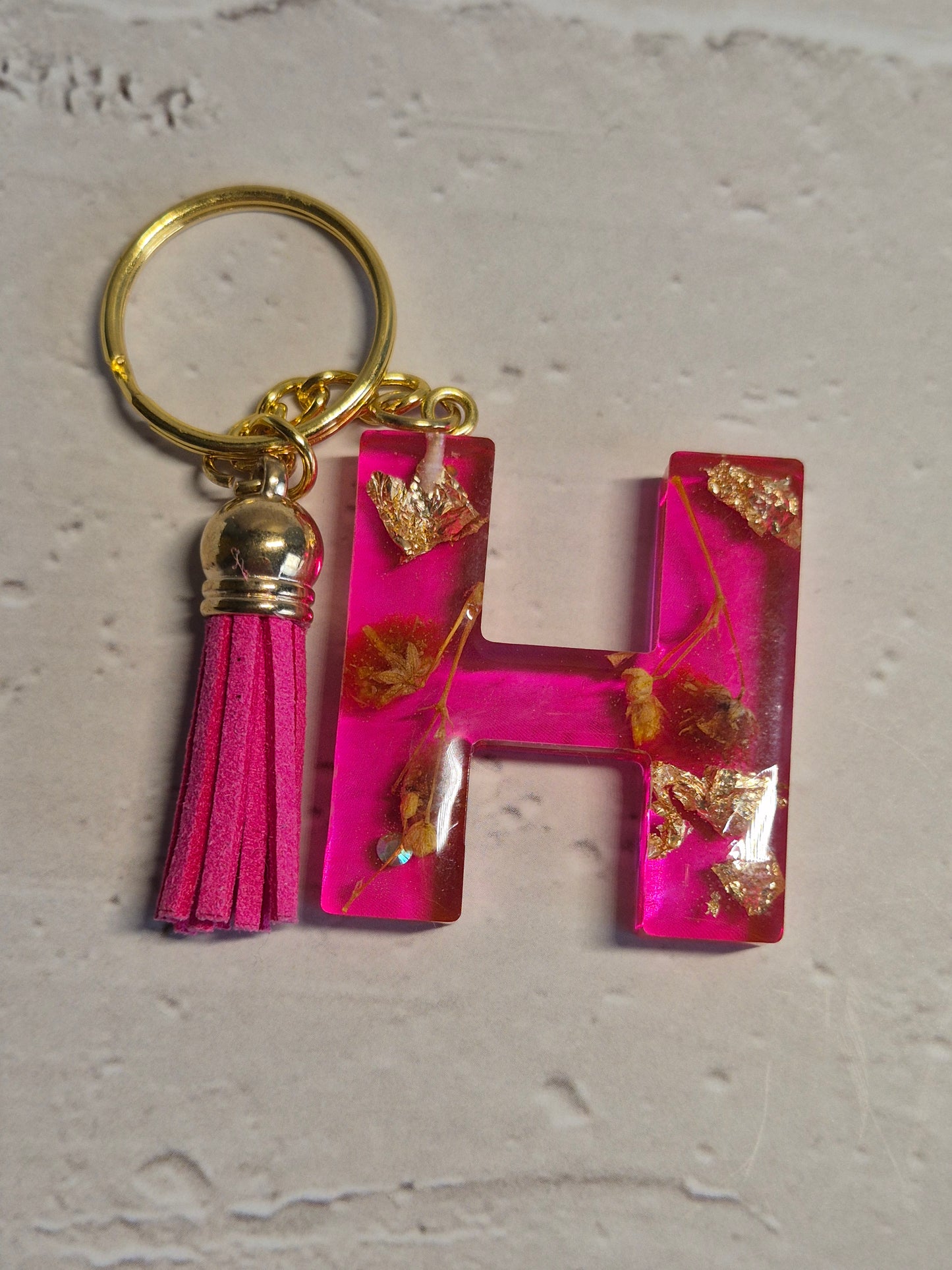 Llavero letra H de resina epoxi fucsia transparente con flores secas, pan de oro y borla fucsia